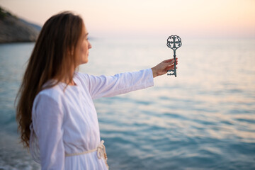 A girl on the seashore in a white dress holds an ancient key in her hands.