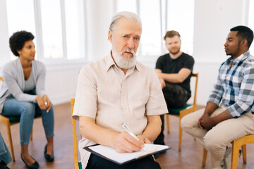 Medium shot portrait of gray-haired mature adult man posing looking at camera while group of multiethnic people sitting in circle talking in background during therapy session. Concept of mental health