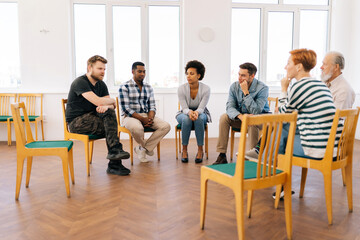 Portrait of group therapy session with diverse people sharing sad problems and stories during psychotherapy session. Multi-ethnic men and women sitting in circle talking about mental health issues.