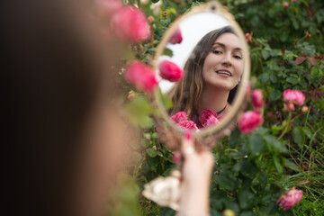 Obraz na płótnie Canvas a beautiful girl in a pink dress in a garden with red roses looks in the mirror