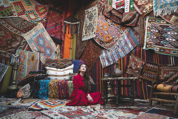 a beautiful girl in a red dress in a traditional turkish interior with many carpets on the wall - obrazy, fototapety, plakaty