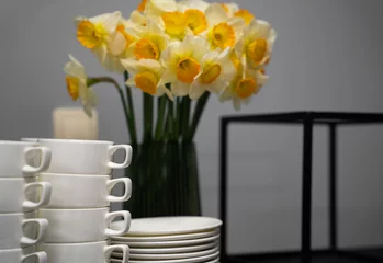 Poster Many clean white cups and plates on the table near the coffee maker and vase with daffodils © Tsyb Oleh