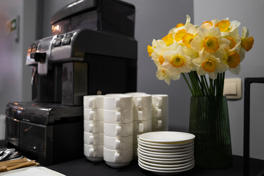 Many clean white cups and plates on the table near the coffee maker and vase with daffodils