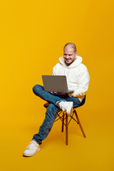 Caucasian handsome cheerful young man working on laptop and sitting on chair, isolated over yellow background