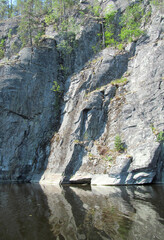 Rocky shore with large stones and trees reflected in calm water of northern lake. Beautiful summer nature of Ladoga lake. Karelia. Russia.