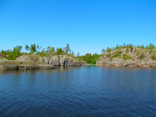 Rocky coast with large stones, trees and reflection in quiet water surface of northern lake. Northern nature and blue sky in summer. Ladoga lake, Karelia, Russia.