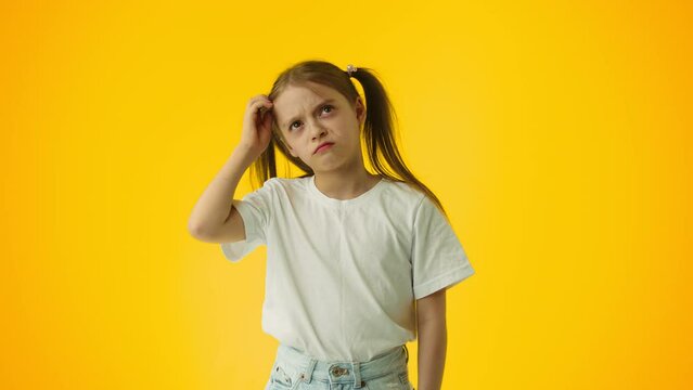 Portrait of a kid girl gesturing with raised hand touches her head, being buffled and confused, posing over yellow color background wall in studio