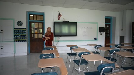 Sad unhappy teacher wearing headscarf in an empty classroom with the lights turned off with US American flag during a pandemic wondering about the future of education.
