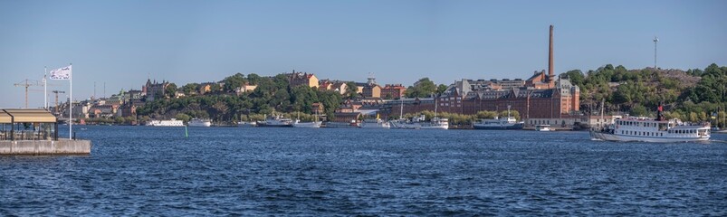 Fototapeta na wymiar Panorama, tourist steam boat and modern ferries, industrial brick buildings, , hill Maria. district Södermalm, moored old fishing and hotel boats, a sunny summer evening in Stockholm