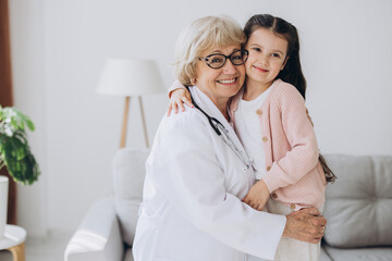Fototapeta premium Portrait of senior nurse wearing white coat, embracing shoulders of happy little preschool patient. Smiling small girl holding favorite toy, posing for photo with attractive general practitioner.