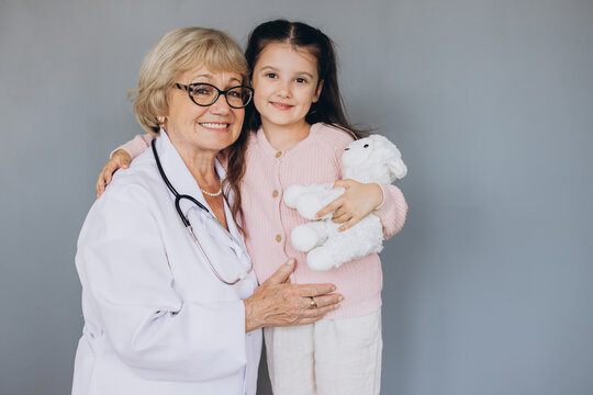 Portrait of senior nurse wearing white coat, embracing shoulders of happy little preschool patient. Smiling small girl holding favorite toy, posing for photo with attractive general practitioner.
