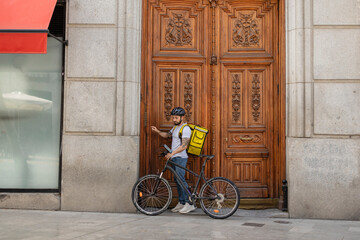 Latino adult man working as a delivery man arrives at his destination to deliver a package on his bicycle with his backpack - obrazy, fototapety, plakaty