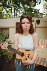Happy hippie girl is having a good time with playing on guitar in camper trailer. Holiday, vacation, trip concept.