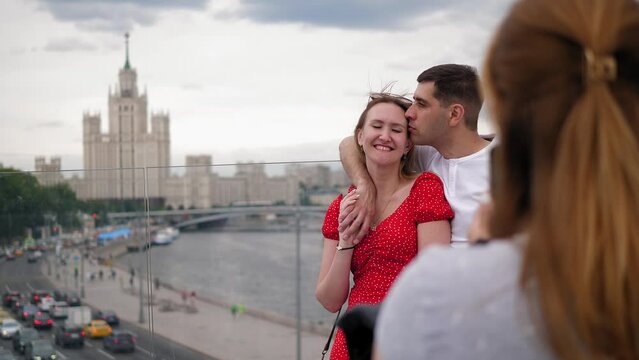 Close-up of a young woman taking pictures on her phone of a couple in love, they are standing on a glass bridge against the background of a river and a big city.