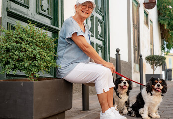 Active senior smiling woman with hat sitting in the street with her two cavalier king charles dogs. Mature lady and her best friends