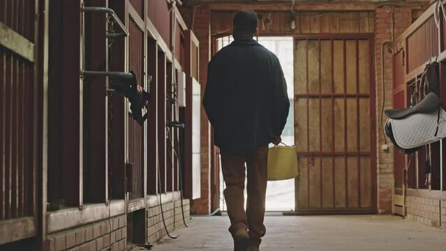 Back view of young African American man walking with bag through horse stable and opening stall gate while working on farm