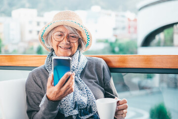 Portrait of relaxed senior woman smiling drinking hot coffee or tea outdoor in cafeteria looking at smartphone