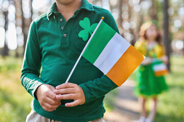 Young boy holding Irish flag in hands on a St Patrick's Day holiday. Unrecognizable kid dressed in...