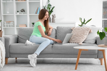 Pretty young woman sitting on grey sofa and using modern laptop in interior of living room