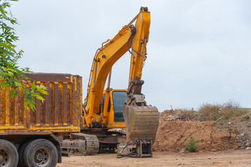 Yellow excavator in a landfill with garbage and soil next to a truck ready for loading.