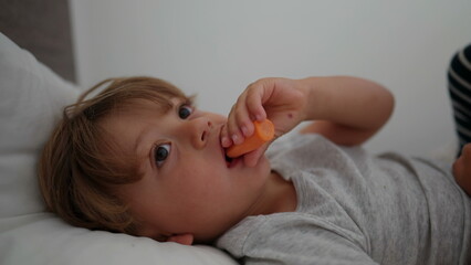 Obraz na płótnie Canvas Little boy eating carrot lying down in bed eating healthy snack