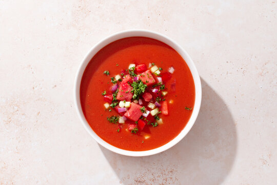 Spanish tomato and watermelon gazpacho cold soup styled and decorated in white plate with ingredients on concrete table in hard light with shadows, top view