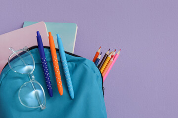 Blue school backpack with different stationery and eyeglasses on purple background