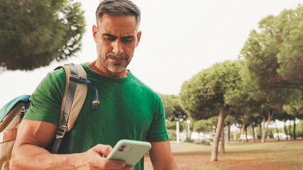 Fototapeta premium Mature man wearing casual clothes, with backpack on his shoulder, stands in the park, sends SMS from mobile phone, always online