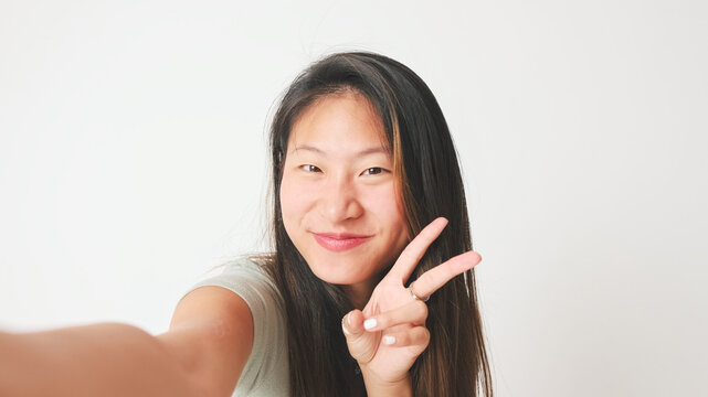 Smiling girl in gray top takes selfie and shows hand victorie, makes video call isolated on white background, studio shot