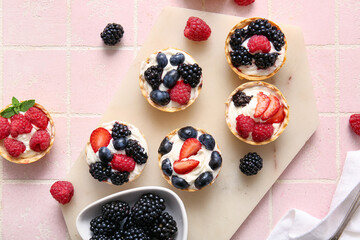Board of tasty tartlets with whipped cream and berries on pink tile background