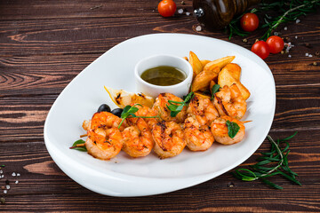 Fried shrimp on a skewer with rustic potatoes, lemon, tomatoes, olives and sauce on a wooden table.
