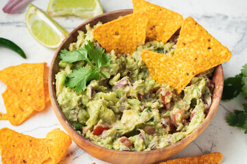 White bowl of traditional Mexican guacamole with nachos on white marble background. Tortilla chips with guacamole sauce dip and ingredients: avocado, cilantro, onion, hot pepper and lime