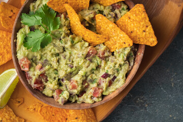 Wooden bowl of traditional Mexican guacamole with nachos on grey concrete background. Tortilla chips with guacamole sauce dip and ingredients: avocado, cilantro, onion, hot pepper and lime. Flat lay
