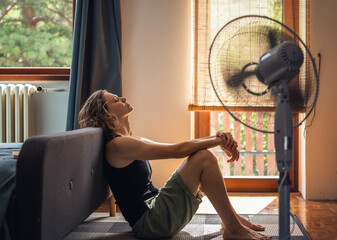 Young woman sitting at home on the floor in front of a fan saving from heat