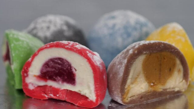 Six cut halves of mochi cakes in brown, green, red, black, yellow and black colors spin while lying on a silver surface. Presentation of flavors on a light background.