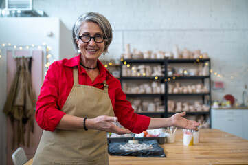 Female ceramist against shelves of handmade ceramics showing welcome gesture inviting to workshop