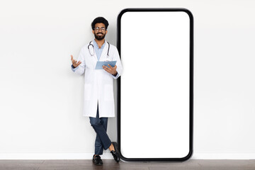 Positive eastern doctor standing by huge smartphone with white screen