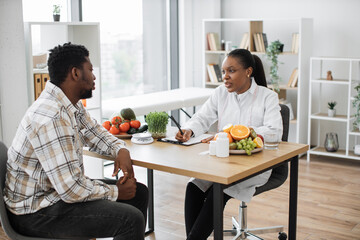 Confident female dietitian in lab coat consulting male patient in doctor's office of clinic....