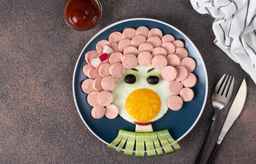 Fun Food for kids - smiling face from fried eggs, sausages, olives and cucumber on blue plate. Creative healthy breakfast for children