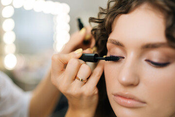 Close-up face of attractive young woman getting applying mascara on eyelashes from makeup artist in beauty salon on blurred background of mirror with bokeh lighting. Concept of fashion and beauty.