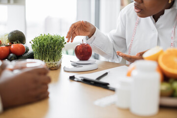 Cropped view of female in doctor's coat weighing fresh apple on digital scale during consultation in hospital. Multiracial nutritionist giving advice on improving diet with low-calorie products.