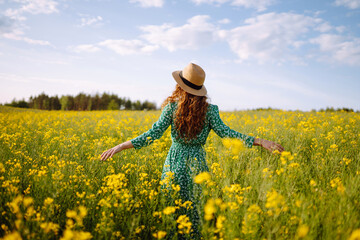 Beautiful woman in bright dress and elegant hat walks and has fun in rapeseed field. Smiling female tourist walking through flowering field, touching yellow flowers. Nature, rest. Summer landscape.