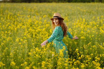 Beautiful woman in bright dress and elegant hat walks and has fun in rapeseed field. Smiling female...
