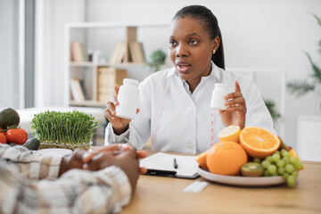 Close up view of multiracial lady in lab coat holding two bottles with capsules during working day in clinic. Pensive expert in diet explaining difference between vitamins and minerals in office.