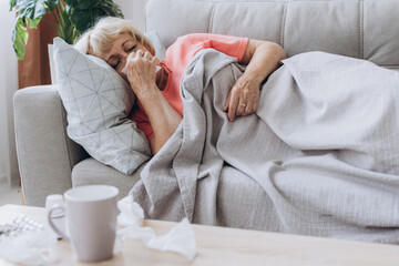 Senior woman being sick having flu lying on sofa. Sick older lady lying in bed with high fever. - obrazy, fototapety, plakaty