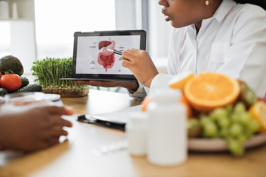 Cropped view of multiracial woman in white coat using tablet with picture of gastrointestinal tract during patient's visit. Nutrition professional illustrating system where food being broken down.