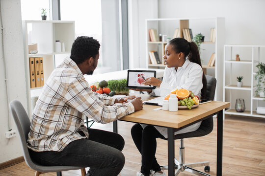 African american specialist in nutrition holding tablet with gastrointestinal tract on screen while counseling man at work. Focused woman explaining client about food intolerance during appointment.