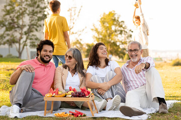 Laughing caucasian multi-generation people have rest, enjoy picnic, food and summer holidays in park, kids play game