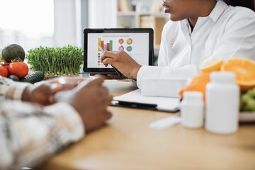 Cropped view of adult lady in lab coat pointing at healthy eating information on device during consultation in hospital. Food expert helping multiethnic patient in developing nutrition and meal plans.