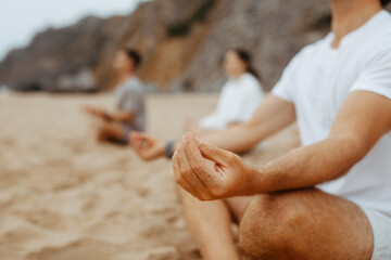 Group of unrecognizable young people meditating together, sitting in lotus position, practising yoga on beach, closeup